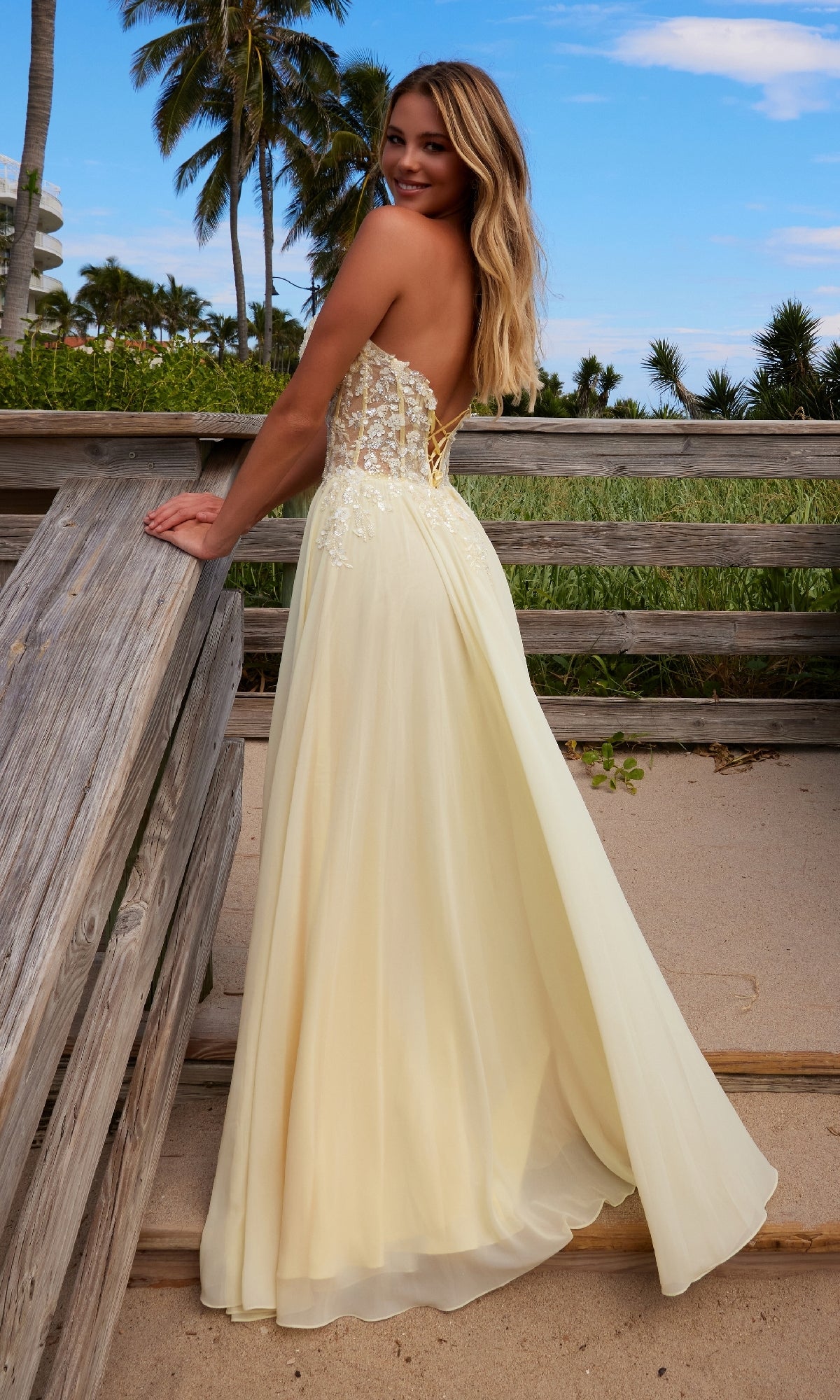 A woman wearing the Nox Anabel C1617 Strapless Sweetheart Prom Dress with a lace-up back stands on a sandy beach near a wooden railing, framed by palm trees and blue sky.